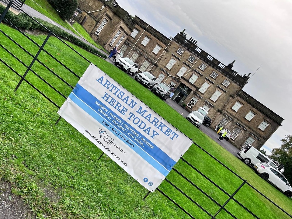 A banner reading "Artisan Market Here Today" hangs on a fence in front of a large historic building with parked cars and people walking on the lawn.